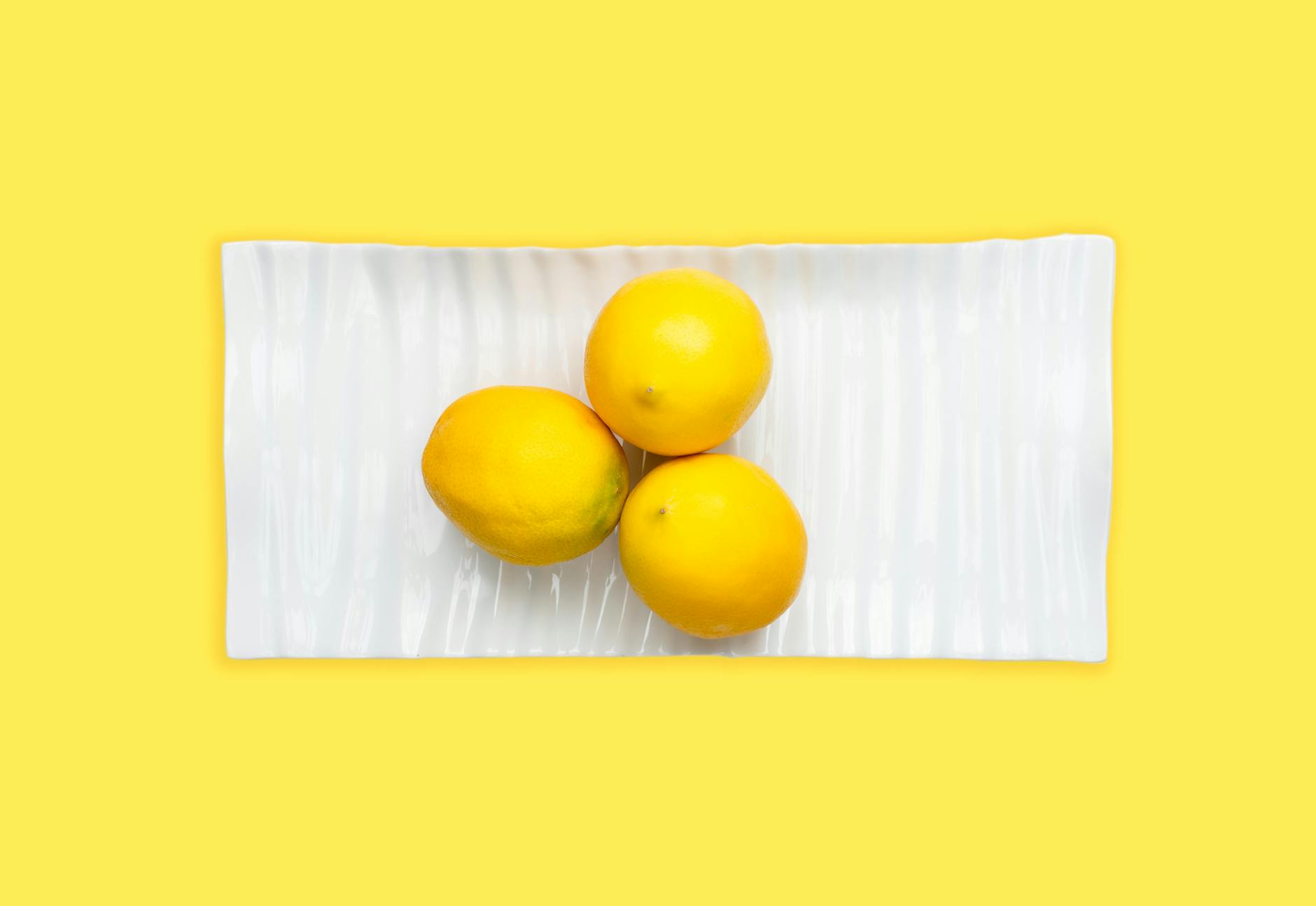 Three fresh lemons arranged on a white plate with vibrant yellow background, symbolizing renewed sensation and citrus freshness