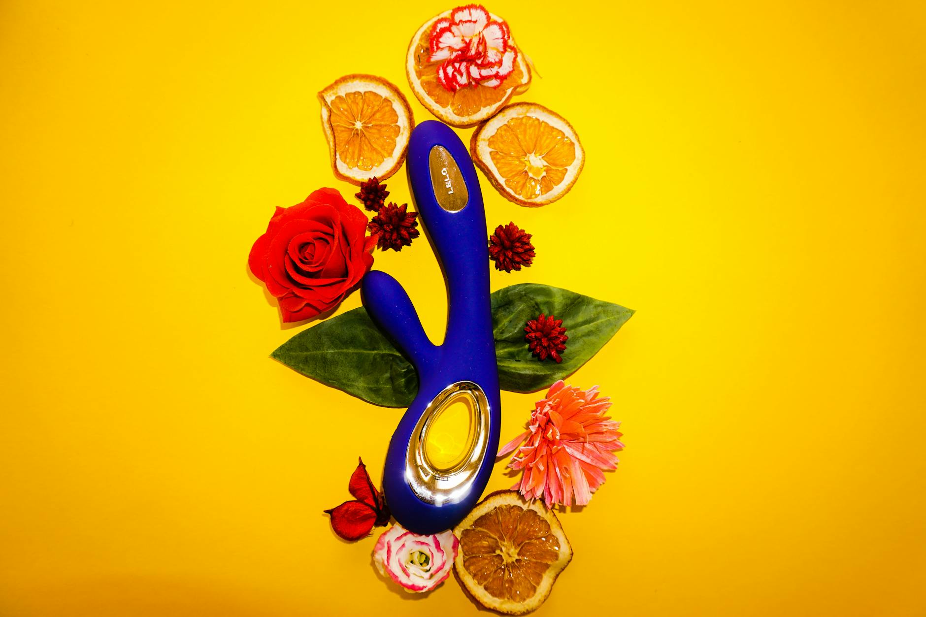 A blue lemon vibrator surrounded by fresh citrus and flowers on a bright yellow surface